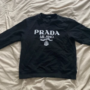 Prada tröja - Svart sweatshirt från Prada Milano med vit logotyp och emblem tryckt på bröstet. Tröjan har rund halsringning och långa ärmar, samt ribbade muddar vid ärmslut och nederkant. Perfekt för en stilren och lyxig look.