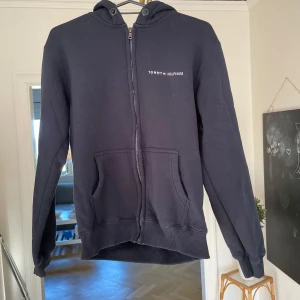 Hoodie - Svart hoodie från Tommy Hilfiger i storlek XS. Klassisk modell med dragkedja framtill, två stora fickor och broderad logga på bröstet. Mjuk insida och huva med snörning gör den perfekt för kyliga dagar.