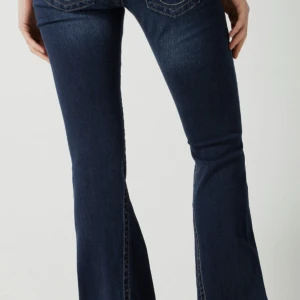 True religion lågmidjade mörkblå jeans - Snygga mörkblå jeans med klassisk bootcut-passform. Jeansen har en figurnära siluett upptill och utsvängda ben nertill. Tillverkade i mjukt denimtyg med diskreta slitningar och kontrastsömmar. Perfekta för en trendig och avslappnad look. Byxorna är i längd 32 men har sytt upp dom lite, men det skulle inte vara några problem få dom som vanliga igen om man spretar upp ändarna igen.