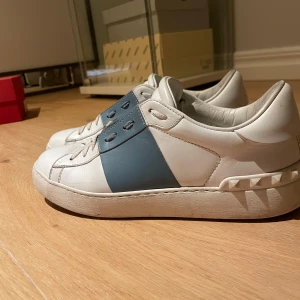 Valentino Garavani Open sneakers vit/blå - Snygga Valentino Garavani Open sneakers i vitt skinn med bred blå läderdetalj över snörningen och ikoniska nitar baktill på sulan. Klassisk rund tå och platt sula. Kommer med original röd box. Perfekta för dig som gillar lyxiga och trendiga sneakers.