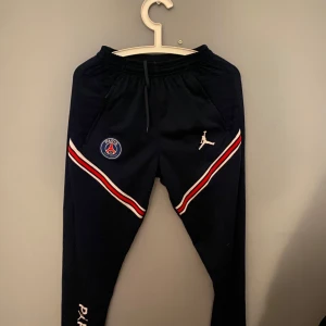 Psg byxor - Snygga svarta träningsbyxor med PSG-logga och ikonisk Jumpman-symbol. Byxorna har röda och vita ränder längs sidorna och elastisk midja med dragsko. Perfekta för sport och avslappnad stil.