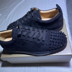 Svarta Christian Louboutin  - Snygga svarta sneakers från Christian Louboutin i mocka med nitar på tån. Skorna har en ikonisk röd sula och snörning. Perfekta för att ge en edgy touch till din stil.