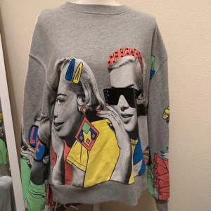 Grå sweatshirt med färgglatt tryck - Cool grå sweatshirt med ett stort, färgglatt tryck av två kvinnor i retrostil. Tröjan har långa ärmar och en avslappnad passform. Perfekt för en unik och stilfull look.