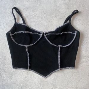 Svart ribbad korsettopp - Snygg svart ribbad korsettopp med kontrasterande vita sömmar. Toppen har smala axelband och en stilren design som framhäver figuren. Perfekt för en trendig look.