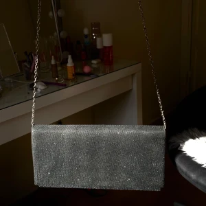 Svart glittrig kuvertväska med kedja - Snygg svart kuvertväska med glittrig yta och en elegant kedja som axelrem. Perfekt för att ge en touch av glamour till din outfit. Väskan har en stilren design och är lätt att matcha med olika stilar.