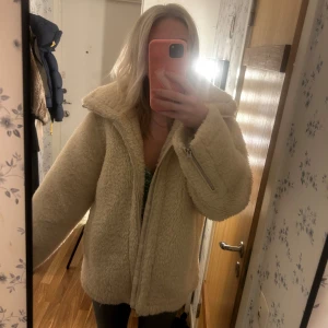 Beige teddyjacka från Arket - Mysig beige teddyjacka från Arket med dragkedja framtill. Perfekt för kyliga dagar med sin fluffiga och varma design. Jackan har en avslappnad passform och långa ärmar, vilket gör den både bekväm och stilren.
