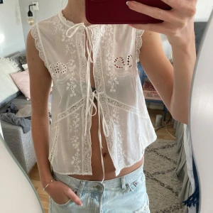 Vit ärmlös blus med spetsdetaljer - Säljer en söt vit ärmlös blus med vackra spetsdetaljer och broderier. Blusen har knytband framtill och en luftig passform, perfekt för varma dagar. Den har en romantisk och bohemisk stil som passar till många olika outfits.