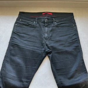 Svarta jeans från Hugo - Snygga svarta jeans från Hugo med klassisk femficksdesign och knappgylf. Byxorna har en stilren look med subtila detaljer och en röd insida vid linningen. Perfekta för en stilren och modern outfit. Scick 8/10 priset är ej hugget i sten