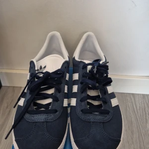 Adidas Gazelle i mörkblå mocka - Snygga Adidas Gazelle sneakers i mörkblå mocka med vita detaljer och snörning. De ikoniska tre ränderna och loggan på hälen ger en klassisk look. Perfekta för en stilren vardagsstil. Helt oanvända och i storlek 37 1/3