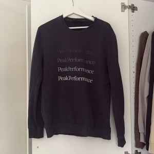 Svart tröja från Peak Performance - Säljer en stilren svart tröja från Peak Performance med deras logga tryckt flera gånger på framsidan. Tröjan har långa ärmar och en rund halsringning. Perfekt för en avslappnad stil.