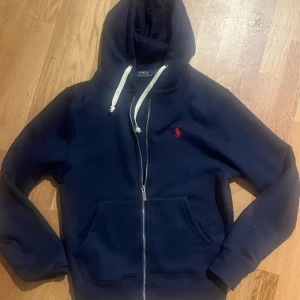 Mörkblå hoodie från Polo Ralph Lauren - Säljer en snygg mörkblå hoodie från Polo Ralph Lauren med dragkedja och vit dragsko. Den har en klassisk design med det ikoniska röda logotypen på bröstet. Perfekt för en avslappnad stil. Pris går såklart att diskutera 