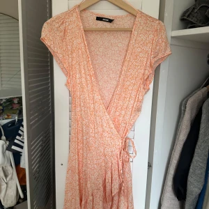 Orange blommig omlottklänning från Bik Bok - Säljer en söt orange omlottklänning från Bik Bok med blommigt mönster. Klänningen har korta ärmar och knytning i midjan. Perfekt för varma dagar!