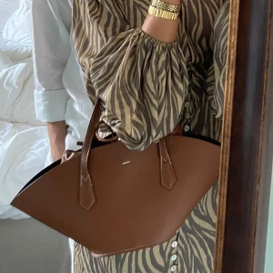 Brun axelväska i skinn  - Snygg brun axelväska i skinn med unik form och stilrena handtag. Väskan har en rymlig insida med ljus foder och praktiska detaljer. Perfekt för att bära med sig alla nödvändigheter med stil. Väskan är från Zara och använd Max 3 gånger