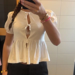 Vit blus med knytband -  vit blus med korta ärmar och knytband framtill. Den har en luftig passform och en söt volangdetalj vid nederkanten. Perfekt för en somrig look!