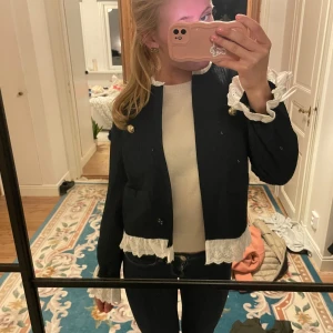 Svart kavaj med spetsdetaljer - Snygg svart jacka med vita spetsdetaljer vid ärmslut och nederkant. Jackan har en elegant design med guldknappar framtill. Perfekt för att ge en stilren touch till din outfit.