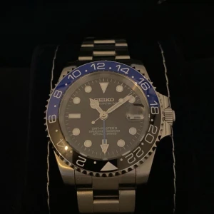 Seiko Batman GMT - Säljer en Seiko klocka med en stilren design. Den har en blå och svart bezel med GMT-funktion och en robust länkarmband i silverfärgat stål. Perfekt för den som gillar en klassisk och funktionell stil.