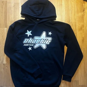 Svart hoodie med stjärnmotiv - Säljer en svart hoodie med ett coolt stjärnmotiv och texten 'chaotic system' på framsidan. Tröjan har en klassisk huva och långa ärmar, perfekt för en avslappnad stil. Passar bra till både jeans och joggers.