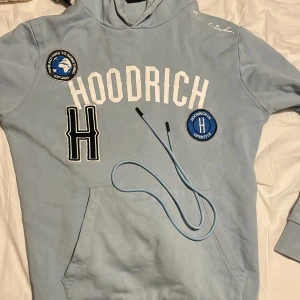 Ljusblå hoodie från Hoodrich - Säljer en ljusblå hoodie från Hoodrich med tryck och broderade detaljer. Den har en stor logga på framsidan och en justerbar huva med snören. Perfekt för en avslappnad stil.
