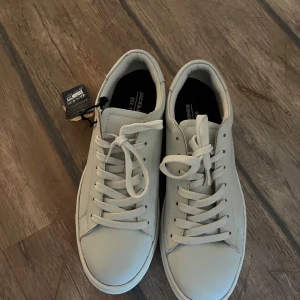 Vita sneakers från Jack & Jones - Snygga vita sneakers från Jack & Jones med klassisk design och snörning. Perfekta för en stilren look. Skorna har en bekväm passform och är tillverkade i syntetmaterial. Nypris 1000, mitt pris 500