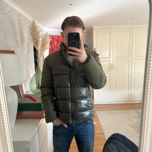 Grön pufferjacka från invicta - Säljer en snygg grön pufferjacka från invicta. Jackan har en glansig finish med quiltade paneler och en praktisk huva. Den är långärmad och har dragkedja.