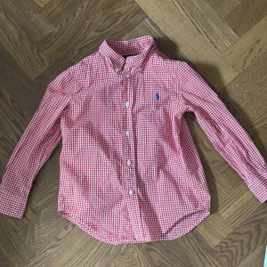 Röd vit rutig skjorta från Ralph Lauren - Snygg röd och vit rutig skjorta från Ralph Lauren med klassisk krage och knappar framtill. Perfekt för en stilren look. Den har långa ärmar och en liten broderad logga på bröstet.