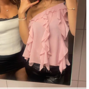 Rosa oneshoulder topp med volanger - Säljer en söt rosa oneshoulder topp med volanger. Perfekt för en trendig look. Toppen har en luftig passform och är gjord i ett lätt material.