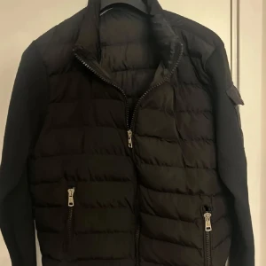 Svart dunjacka från Moncler - Säljer en snygg svart dunjacka från Moncler med quiltad design och dragkedja framtill. Jackan har långa ärmar och praktiska fickor med dragkedjor. Perfekt för kyligare dagar.