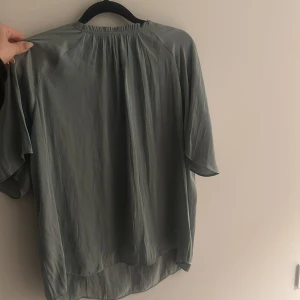 Grön blus från Wera - Säljer en elegant grön blus från Wera. Blusen har en lös passform med korta ärmar och en v-ringad design med knytning vid halsen. Perfekt för en stilren look.