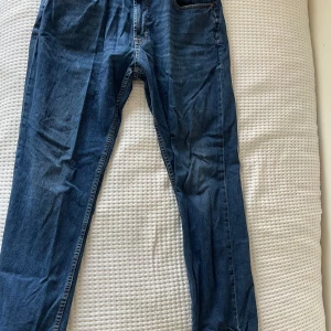 Blå jeansbyxor från Dressmann - Snygga blå jeansbyxor från Dressmann med klassisk design. De har en normal passform och är perfekta för en avslappnad stil. Byxorna har fickor både fram och bak samt en knapp och dragkedja i gylfen.