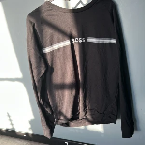 Svart tröja från BOSS - Säljer en svart tröja från BOSS Homewear med vita ränder och texten 'BOSS' på framsidan. Tröjan har långa ärmar och en rund halsringning. Perfekt för en avslappnad stil.