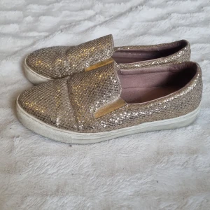 Glittriga slip-on skor - Snygga slip-on skor i glittrigt guld med en bekväm vit sula. I gott skick, lite smutsiga.