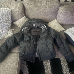 Svart dunjacka från Burberry - Säljer en snygg svart dunjacka från Burberry. Jackan har en quiltad design med dragkedja och en huva med ett stilrent rutigt mönster på insidan. Perfekt för kyliga dagar när du vill hålla stilen. 🖤
