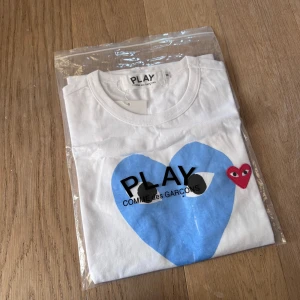 Vit t-shirt från Comme des Garçons - Säljer en vit t-shirt från Comme des Garçons med ett stort blått hjärta och ett litet rött hjärta med ögon på framsidan. T-shirten har korta ärmar och en rund halsringning. Perfekt för en stilren och lekfull look. Tröjan är ny med tags.