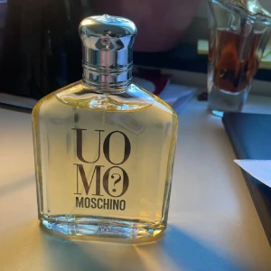 Uomo Moschino Parfym - Säljer en elegant flaska Uomo Moschino parfym. Flaskan har en stilren design med en silverfärgad kork och tydlig logotyp på framsidan. Doften är klassisk och maskulin, perfekt för den som vill ha en tidlös signaturdoft.