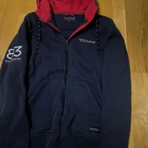 Marinblå hoodie från Race Marine - Säljer en marinblå hoodie från Race Marine med röd insida på huvan och dragkedja framtill. Den har en tryckt logga på bröstet och ärmarna samt snörning i huvan. Perfekt för en avslappnad stil.
