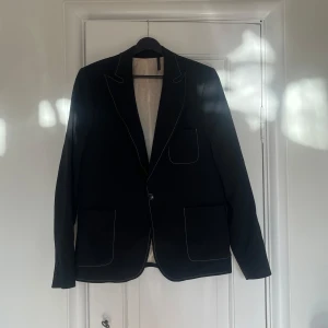 Svart kavaj från Whyred - Snygg svart kavaj från Whyred med klassisk design. Kavajen har två knappar framtill och flera fickor. Perfekt för en stilren look.