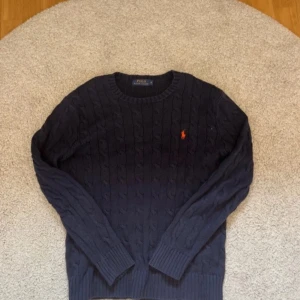 Mörkblå stickad tröja från Ralph Lauren - Säljer en stilren mörkblå stickad tröja från Ralph Lauren med kabelstickat mönster. Tröjan har långa ärmar och en liten broderad logga i orange på bröstet. Perfekt för kyliga dagar!