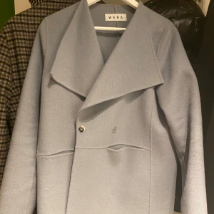 Ljusblå kappa från Wera - Elegant ljusblå kappa från Wera med en stilren design. Kappan har en bred krage och knäpps med knappar framtill. Perfekt för kyligare dagar och ger en sofistikerad look.