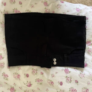 Svart kortkjol  - Snygg svart kortkjol med knappar framtill. Kjolen har en stilren design med fickor baktill och är perfekt för en trendig look.