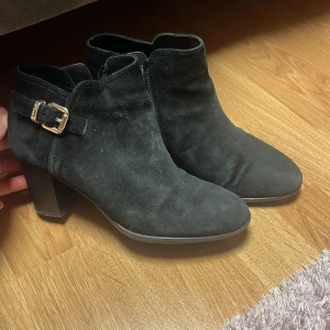 Svarta mocka stövletter med klack och spänne - Säljer ett par svarta stövletter i mocka med bred klack och dekorativt spänne i guld på sidan. Skorna har dragkedja på insidan för enkel påtagning och en rund tå. Perfekta för dig som vill ha en stilren och trendig look.