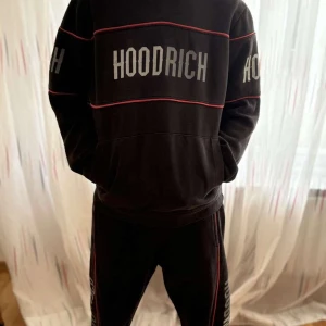 Hoodritch sett, svart hoodie med tryck och svarta byxor med tryck - Hoodric sett, svart  hoodie från Hoodrich med stora vita logotexter på bröst, rygg och ärmar. Hoodien har röda linjer som detaljer och en klassisk huva. Byxor som matchar till hoodien. Hoodritch text på sidorna av byxorna med samma röda linijer. Använd fåtal gånger.