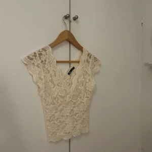 Vit spetsblus från Zara - Säljer en superfin vit spetsblus från Zara med v-ringning och vackra vågiga kanter. Blusen är ärmlös och har ett romantiskt spetsmönster över hela plagget. Perfekt att styla till olika looks!