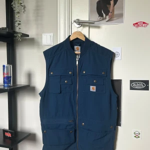 Blå väst från Carhartt med fickor - Säljer en blå väst från Carhartt med flera fickor framtill och dragkedja. Västen har guldiga knappar med Carhartt-logga och klassisk Carhartt-patch på bröstet. Perfekt för lager-på-lager och streetwear.