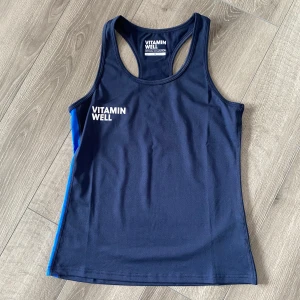 Mörkblå träningstopp från Vitamin Well - Säljer en mörkblå linnetopp från Vitamin Well med vita logotyper fram och bak samt blå sidopanel. Sportig modell med brottarrygg, perfekt för träning. Materialet är syntetiskt och snabbtorkande.