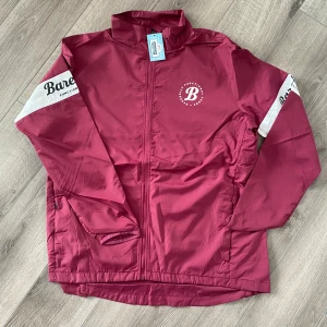 Vinröd vindjacka från Barebells - Säljer en vinröd vindjacka från Barebells Ambassador Collection. Jackan har dragkedja framtill, ståkrage och vita detaljer med logga på ärmarna samt stor logga på ryggen. Perfekt för utomhusaktiviteter och träning.