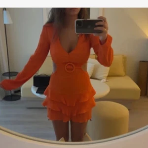 Orange volang klänning - Säljer en orange kortklänning med djup v-ringning, långa ärmar och markerad midja med dekorativt bälte. Kjolen har flera lager volanger för en extra snygg look. Perfekt för dig som vill sticka ut!
