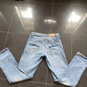 Ljusblå jeans från Nudie Jeans - Säljer ett par klassiska ljusblå jeans från Nudie Jeans med raka ben och femficksdesign. Jeansen har orangea sömmar och Nudie-logga på bakfickan. Perfekta för dig som gillar en tidlös denimlook.