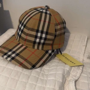 Rutig keps från Burberry - Snygg keps från Burberry i klassiskt beige rutigt mönster med svarta, vita och röda detaljer. Kepsen har böjd skärm och justerbar rem baktill. Perfekt accessoar för en trendig look.
