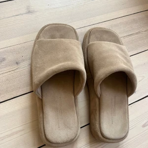 Beige slip-in sandaler med bred rem  - Säljer ett par beige sandaler med bred rem och fyrkantig tå från Arket. De har en platt sula och är tillverkade i mocka-liknande material. Perfekta för sommaren och enkla att glida i.