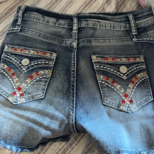 Blå jeansshorts med broderade bakfickor - Säljer ett par blå jeansshorts med fransig kant och snygga broderade detaljer i rött, vitt och beige på bakfickorna. Shortsen har normal passform, klassiska fickor fram och bak samt bälteshällor. Storlek xs/s/m de är töj bara så  de passar även m på helt nya
